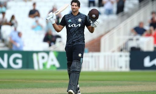 Cricketer wearing a dark-colored uniform holding a bat in one hand and a helmet in the other, acknowledging the crowd after completing a milestone on the field.